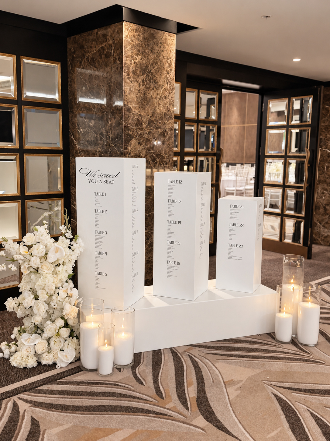 Luxury wedding seating chart display featuring three white personalised seating chart plinths on a white platform, styled with a large white floral arrangement and six glass cylinder candles by Luxury Lane Auckland