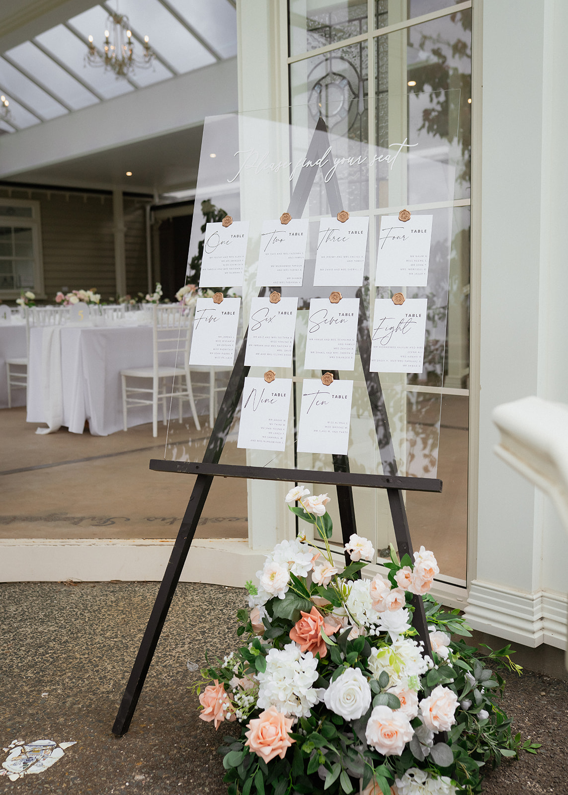 Luxury Lane acrylic wedding seating chart hire in Auckland, featuring personalised table cards with wax seals, black easel and a white and blush floral arrangement.